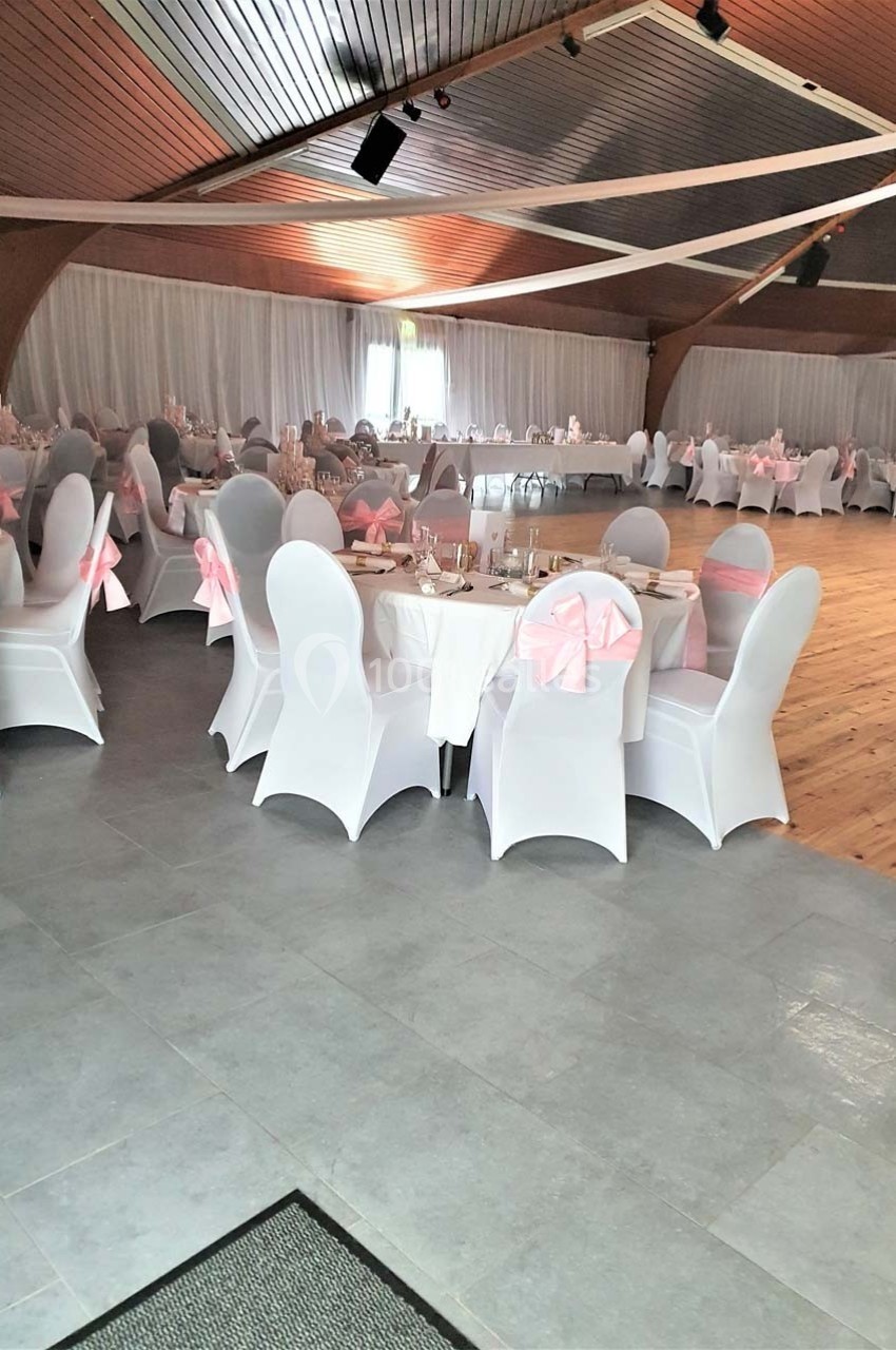 Salle de réception décorée avec des nappes blanches et des nœuds roses sur les chaises, tables dressées pour un événement.