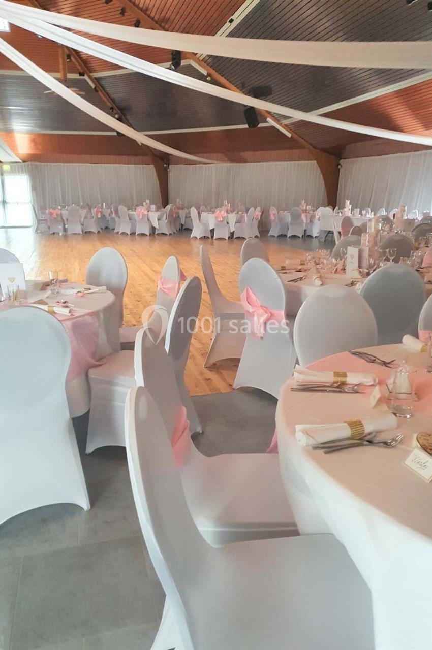 Salle de réception décorée avec des tables rondes, nappes blanches, chaises houssées et rubans roses.