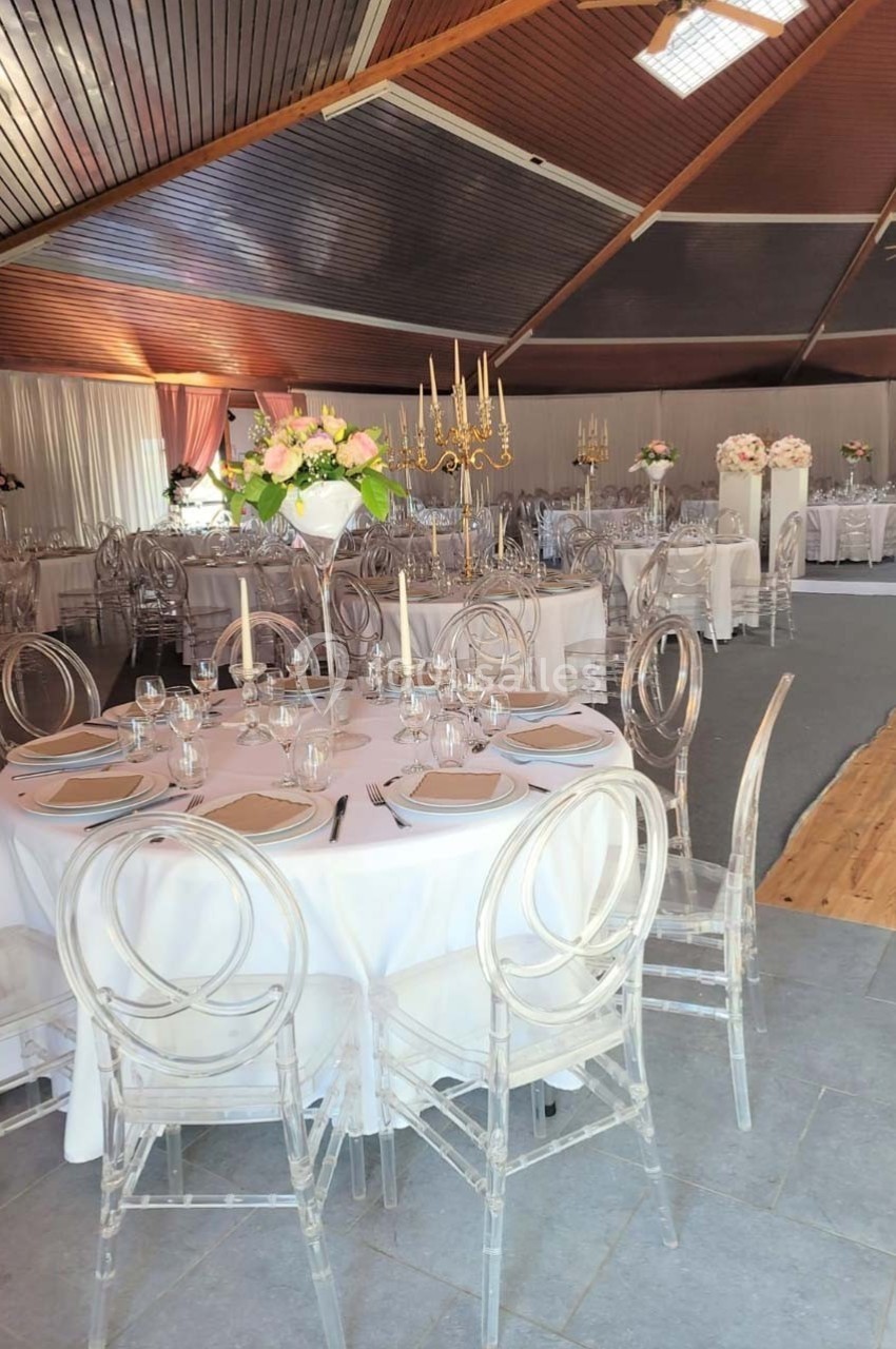 Salle de réception décorée avec des tables rondes, nappes blanches, chaises transparentes et centres de table fleuris.