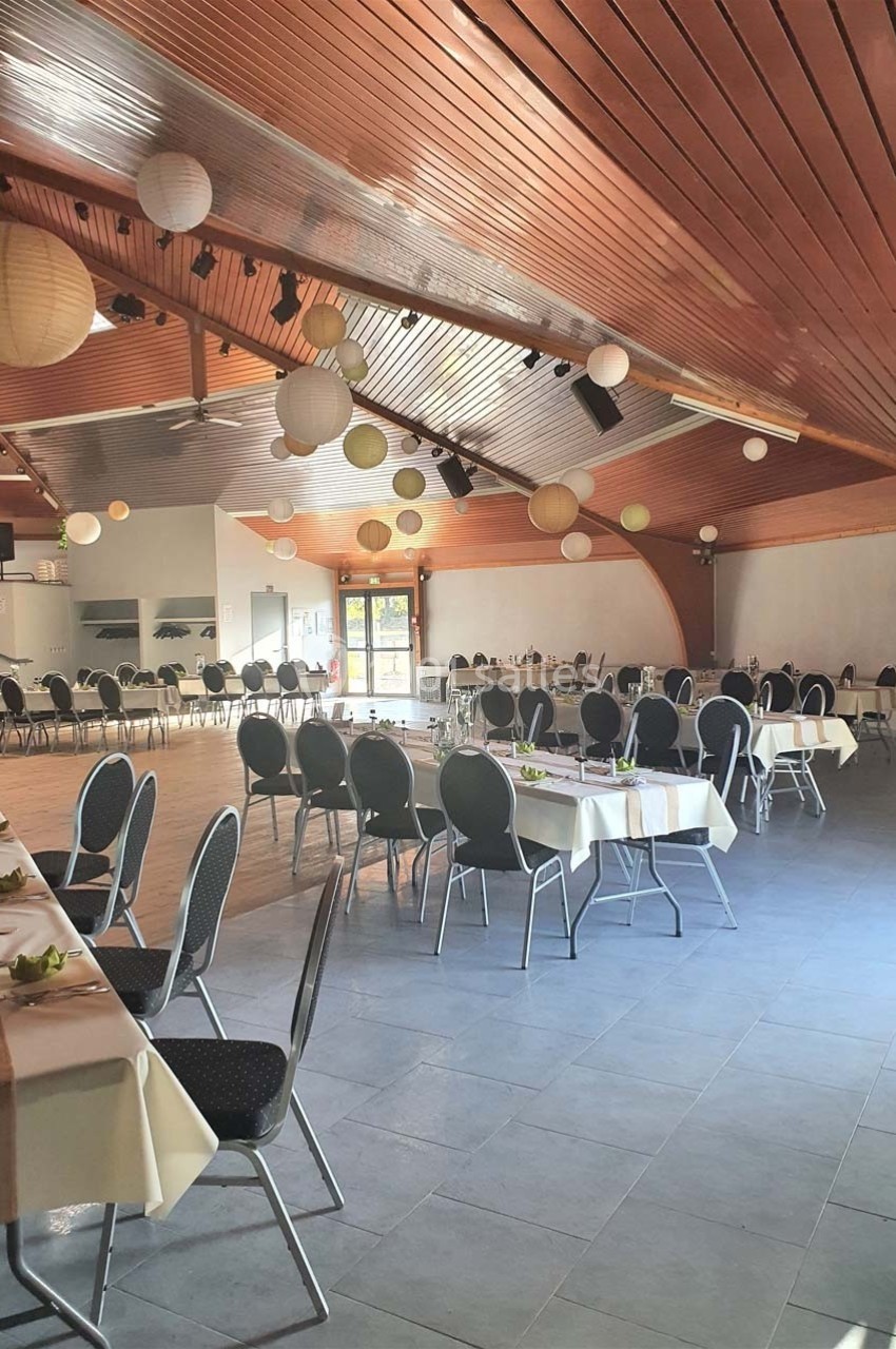 Salle de réception lumineuse avec tables dressées, chaises noires et décorations suspendues sous un plafond en bois.