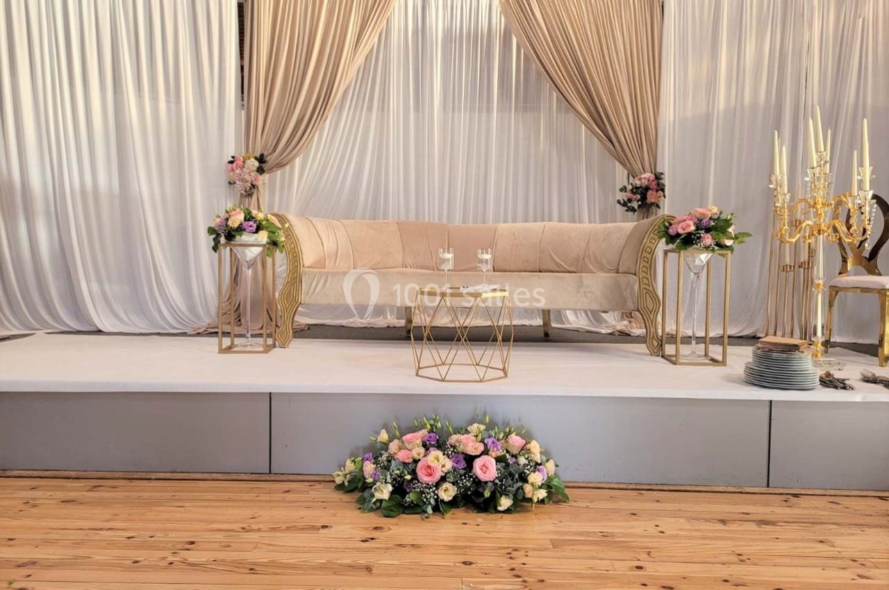 Canapé beige décoré de fleurs pastel, entouré de rideaux dorés et blancs, sur une estrade en bois.