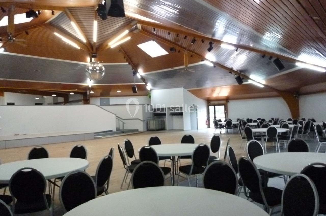 Salle spacieuse avec plafond en bois, tables rondes et chaises noires, éclairage suspendu et scène en fond.