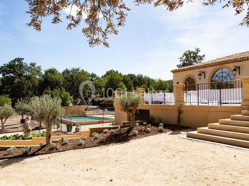 Maison avec terrasse surélevée, jardin paysager, piscine clôturée et arbres sous un ciel dégagé.