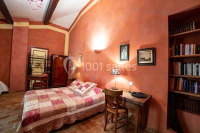 Chambre lumineuse avec murs orange, lit double, sol en terre cuite, table près d'une fenêtre et cloison en bois.