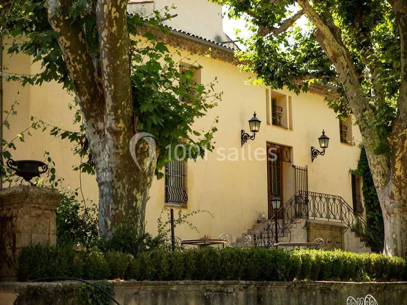 Façade d'une maison ancienne entourée d'arbres et de verdure, avec une terrasse et des lampadaires en fer forgé.