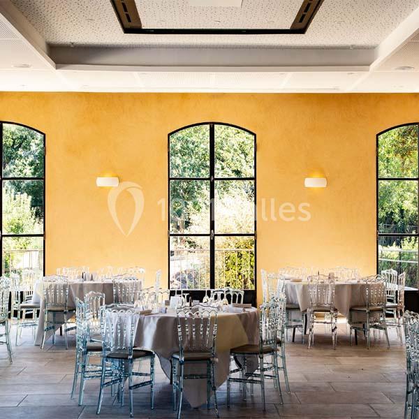 Salle lumineuse avec murs jaunes, grandes fenêtres cintrées, tables rondes dressées et chaises transparentes.