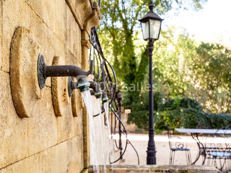 Fontaine en pierre avec plusieurs robinets d'où s'écoule de l'eau, située dans un jardin avec mobilier et lampadaire.
