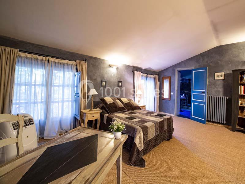 Chambre lumineuse avec lit double, bureau en bois, fenêtres avec rideaux clairs et murs aux tons gris.