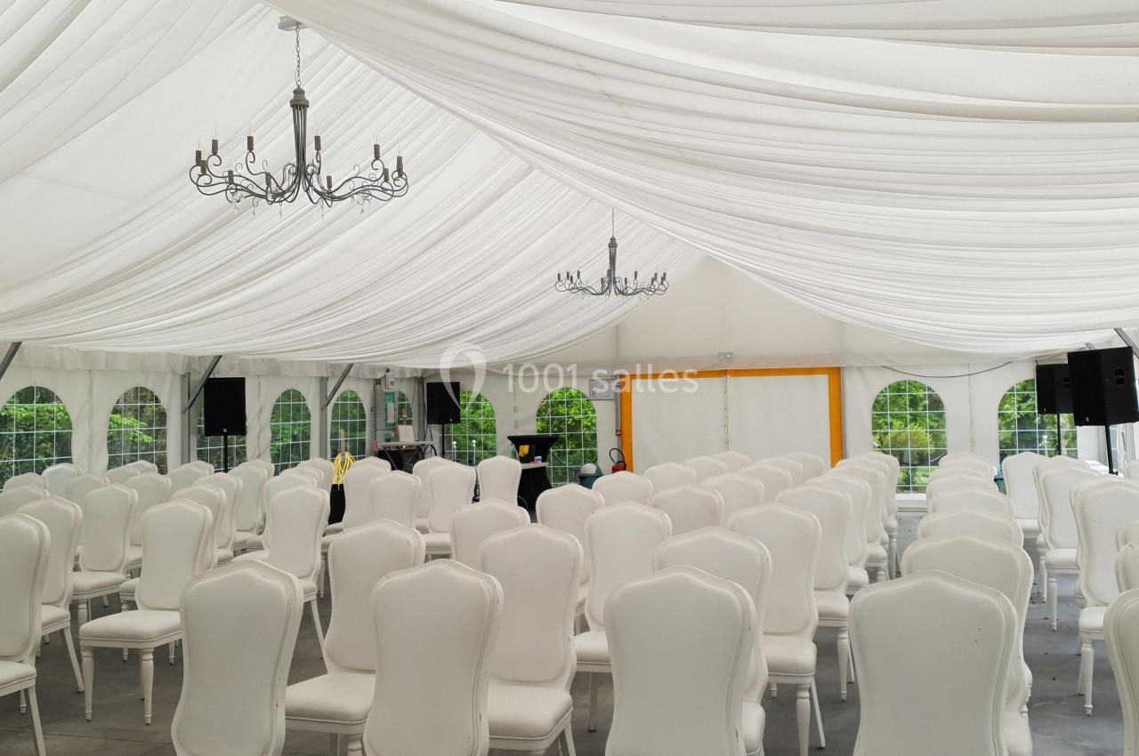 Intérieur d'une tente blanche avec des chaises alignées et des lustres suspendus, préparée pour un événement.