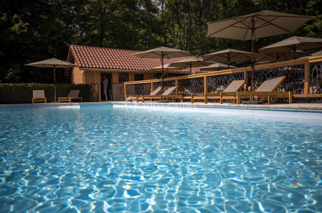 Piscine extérieure entourée de transats en bois et de parasols, avec un bâtiment en bois en arrière-plan.