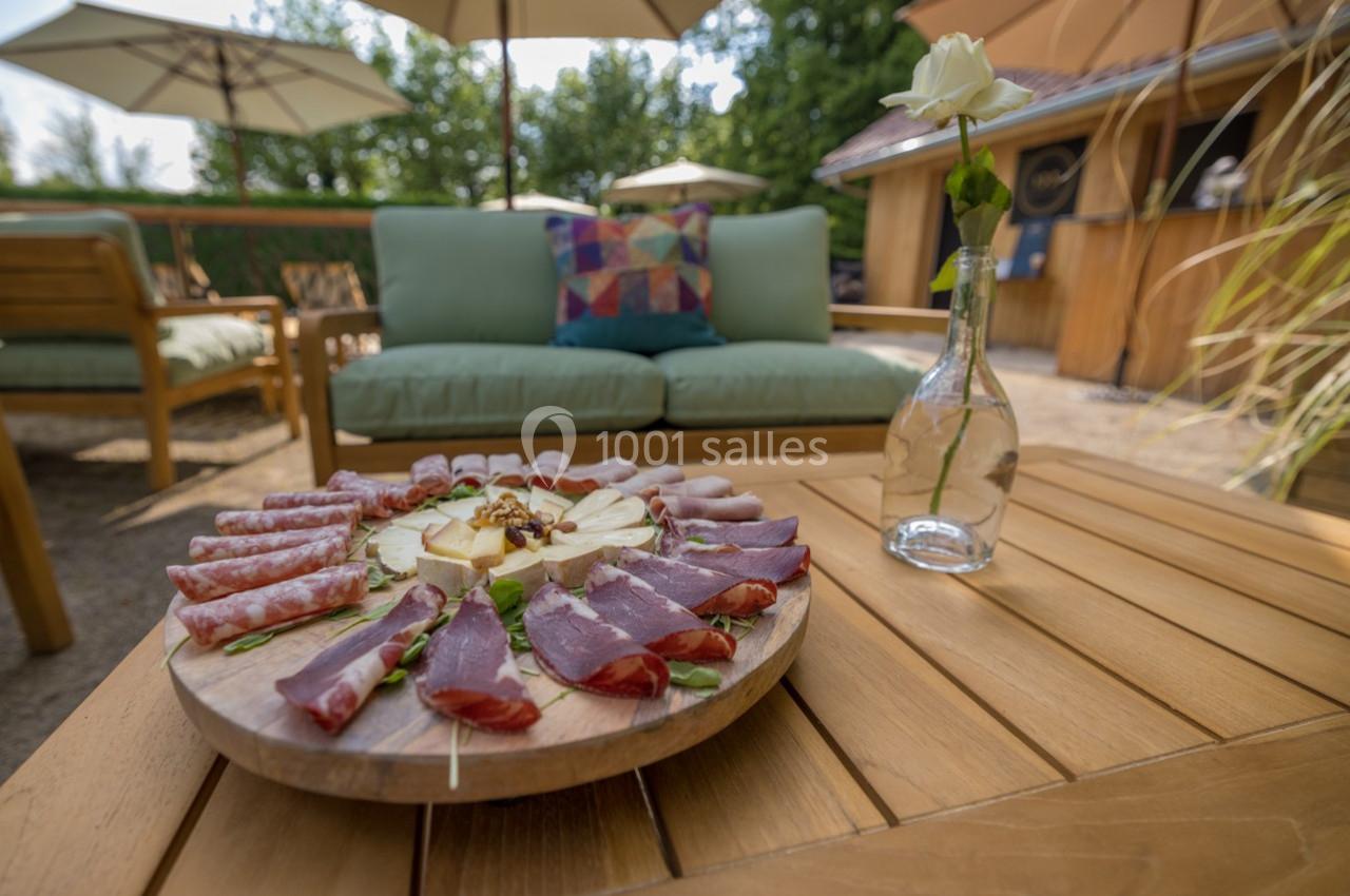 Plateau de charcuterie et fromage sur une table en bois, en terrasse avec mobilier de jardin et parasols en arrière-plan.