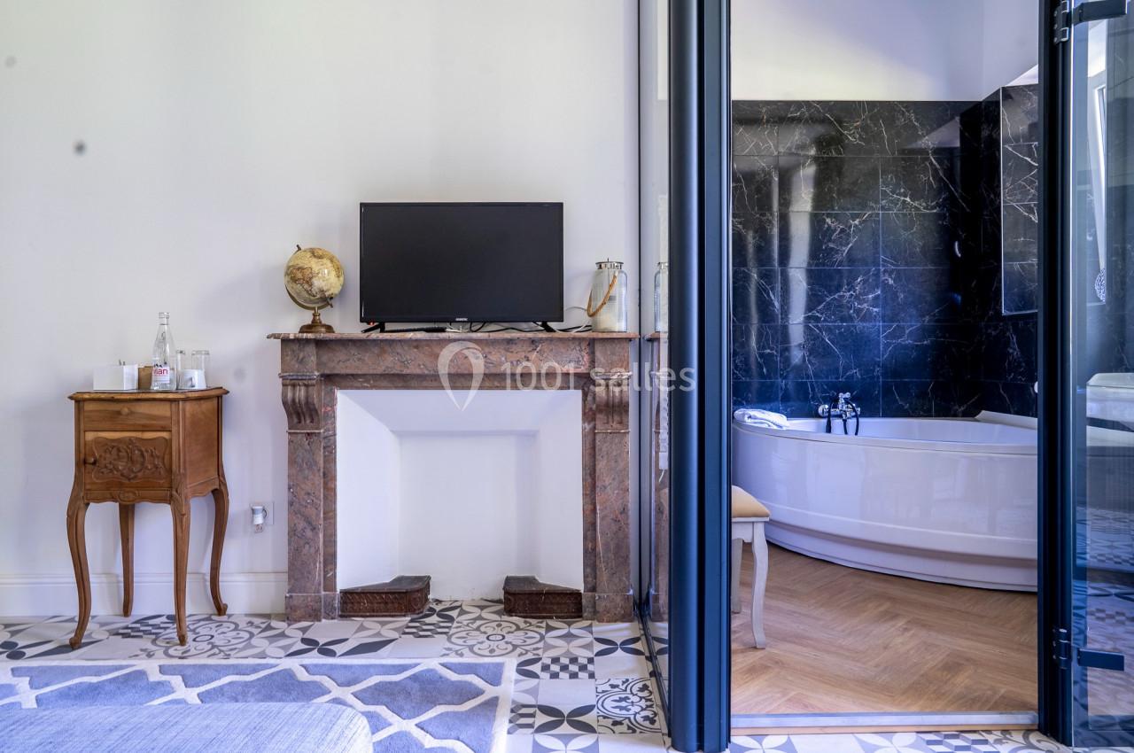 Cheminée en marbre avec télévision, petite table en bois et salle de bain moderne avec baignoire en arrière-plan.