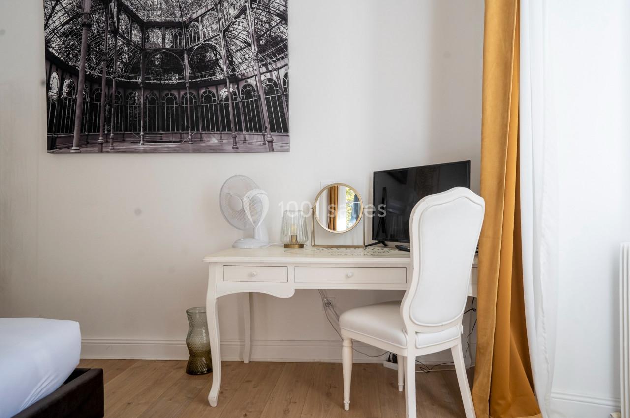 Bureau blanc avec chaise assortie, écran, miroir, ventilateur et décoration, près d'un rideau doré et d'un lit.