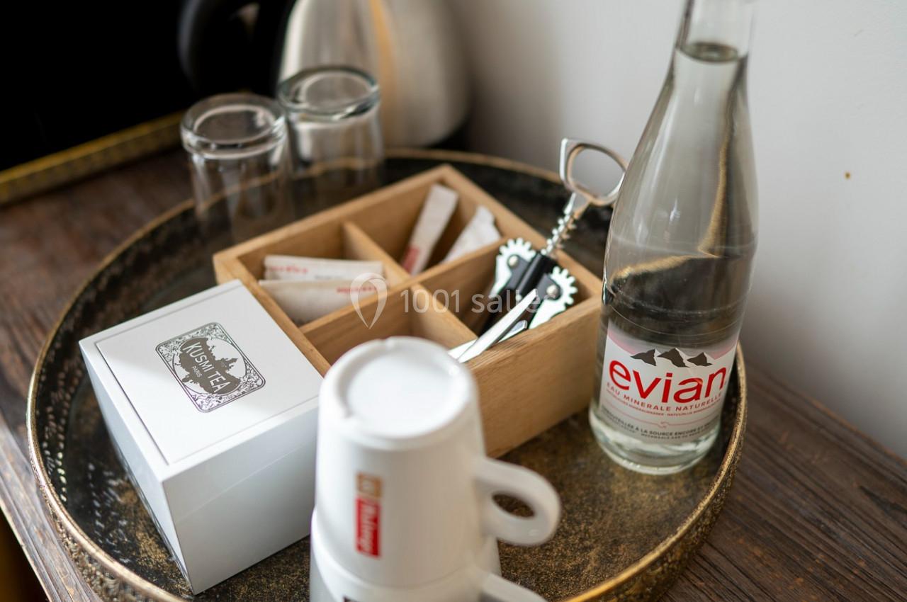 Plateau avec une bouteille d'eau, tasses empilées, sachets de thé, cuillères et une bouilloire en arrière-plan.