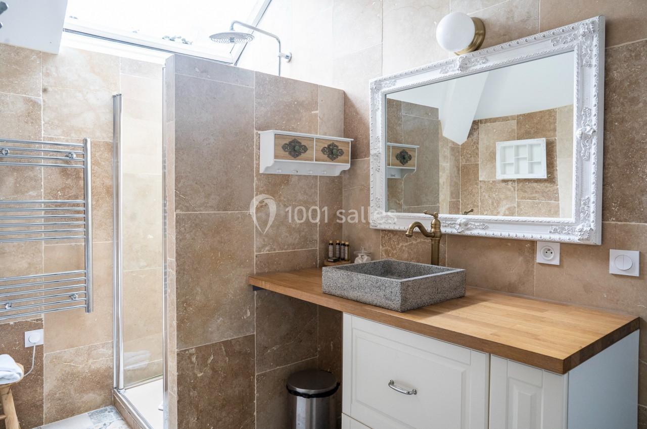 Salle de bain avec douche, lavabo en pierre sur meuble en bois, miroir encadré et carrelage beige.