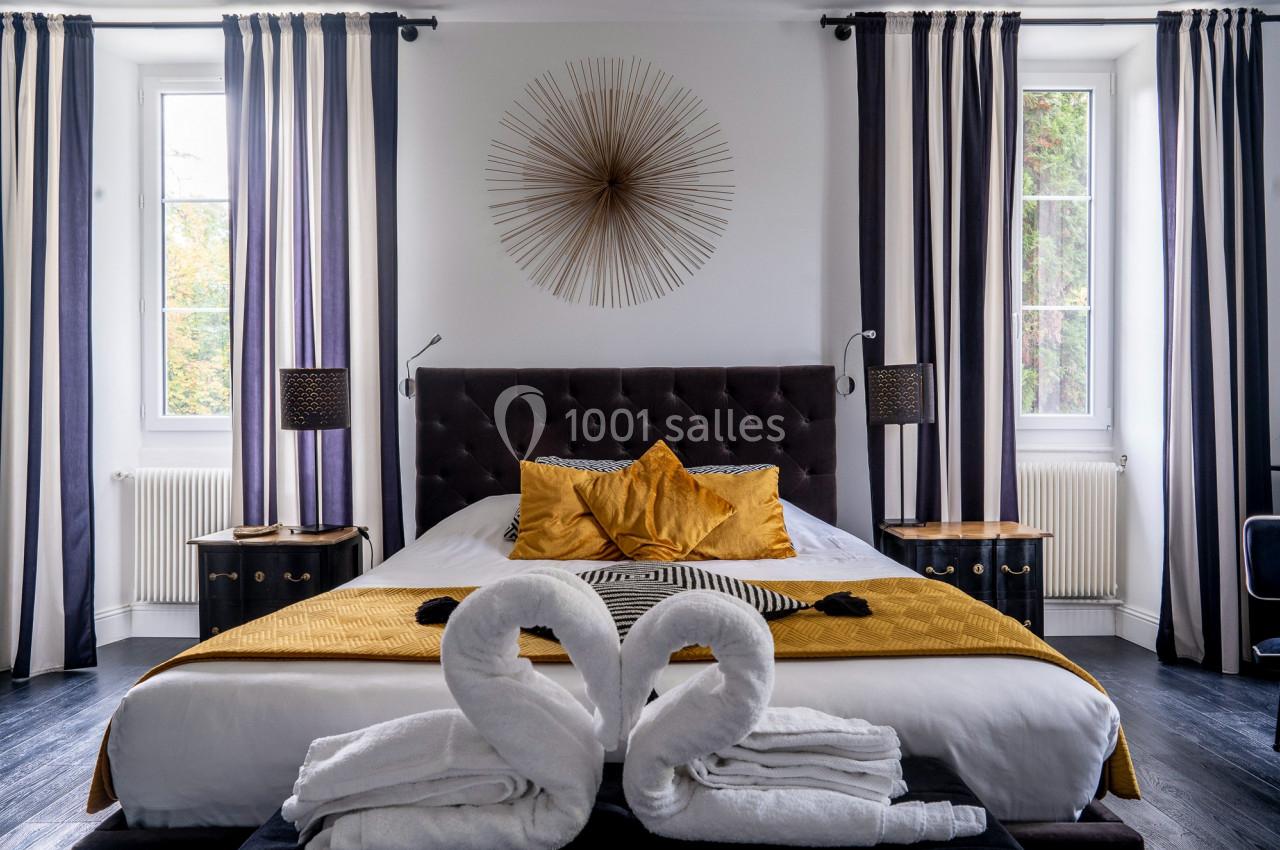 Chambre élégante avec lit double, coussins dorés, serviettes pliées en cygnes et grandes fenêtres encadrées de rideaux rayés.