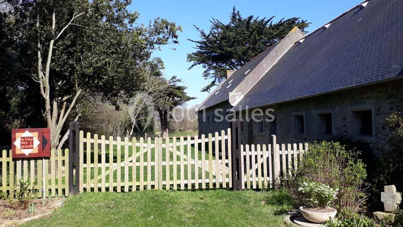 Clôture en bois devant un bâtiment en pierre avec un jardin arboré et un ciel dégagé.