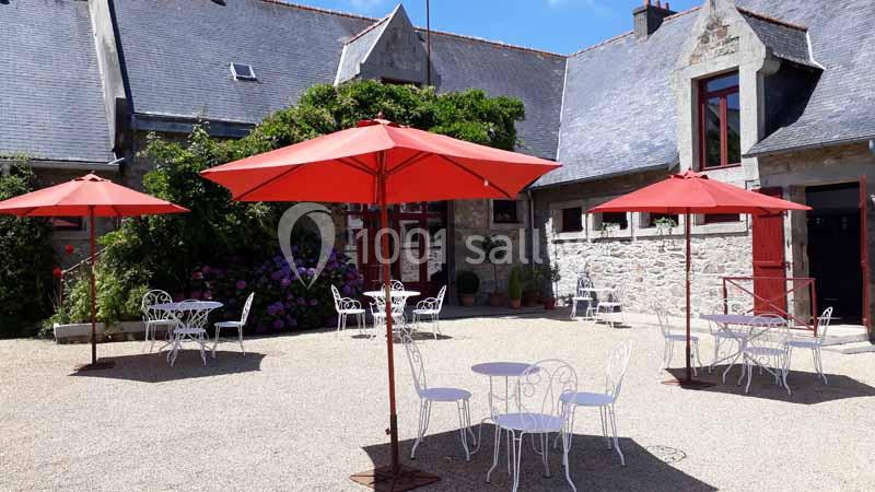 Cour intérieure avec tables et chaises blanches sous des parasols rouges, entourée de bâtiments en pierre.