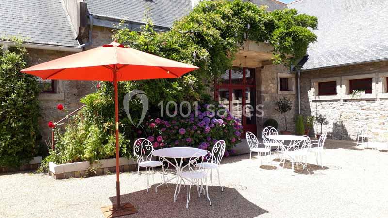 Cour ensoleillée avec tables et chaises blanches en fer forgé, parasol rouge et façade en pierre ornée de végétation.
