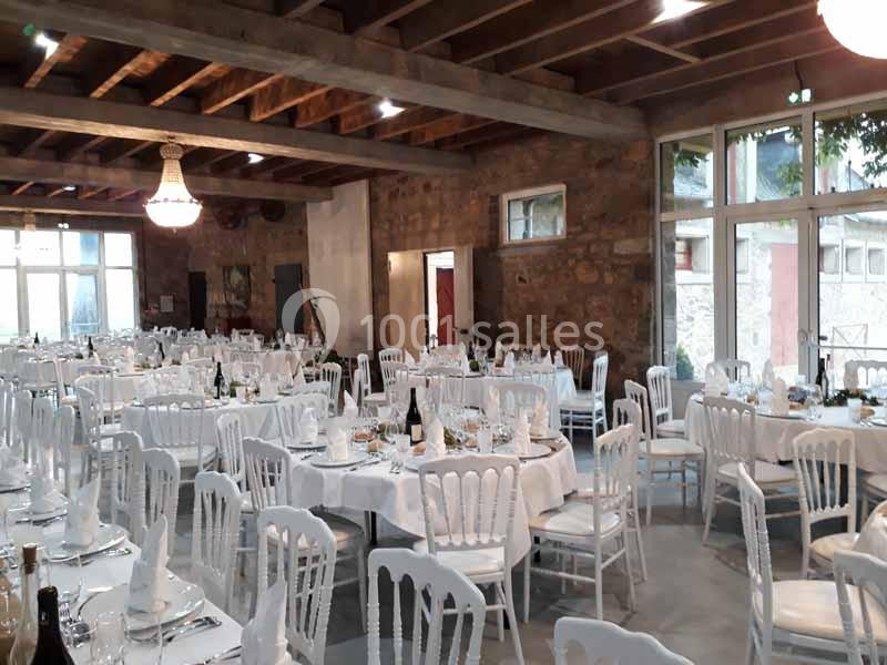 Salle de réception lumineuse avec tables rondes dressées, chaises blanches et poutres apparentes au plafond.
