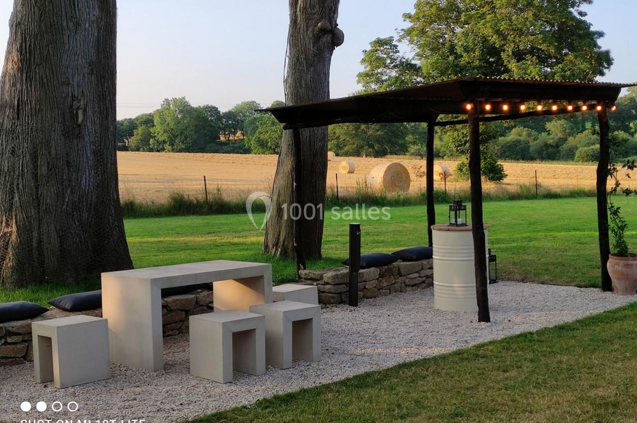 Espace extérieur avec table et bancs modernes sous une pergola éclairée, entouré de pelouse et de champs.