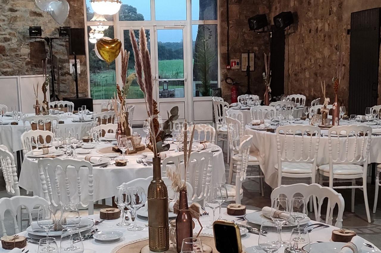 Salle de réception décorée avec des tables rondes, nappes blanches, chaises assorties et ornements champêtres.