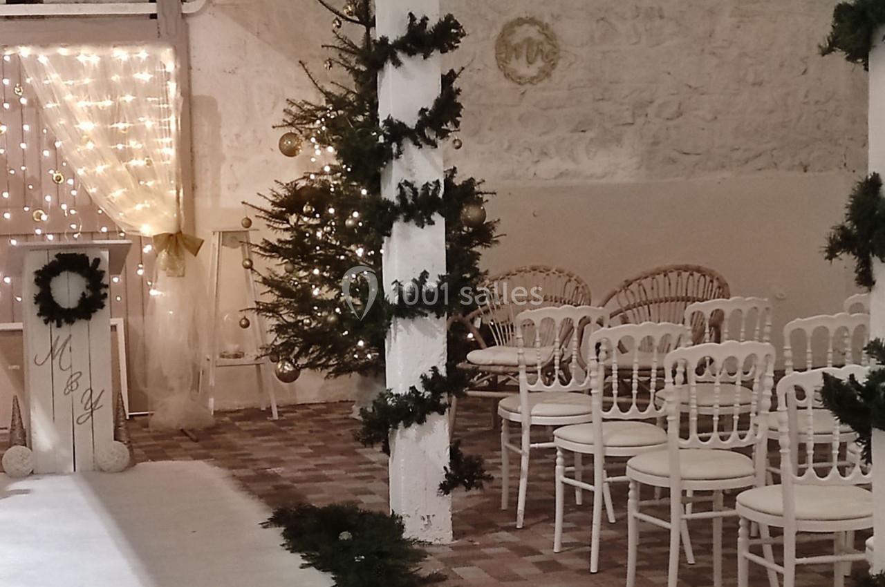 Salle décorée pour une cérémonie avec chaises blanches, sapin illuminé et guirlandes lumineuses sur fond de mur en pierre.