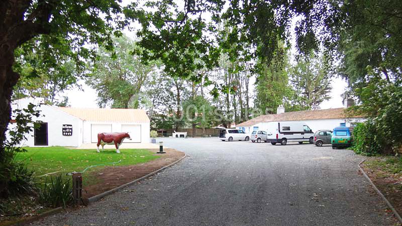 Cour avec bâtiments blancs, cheval en statue, véhicules stationnés et arbres entourant l'espace.