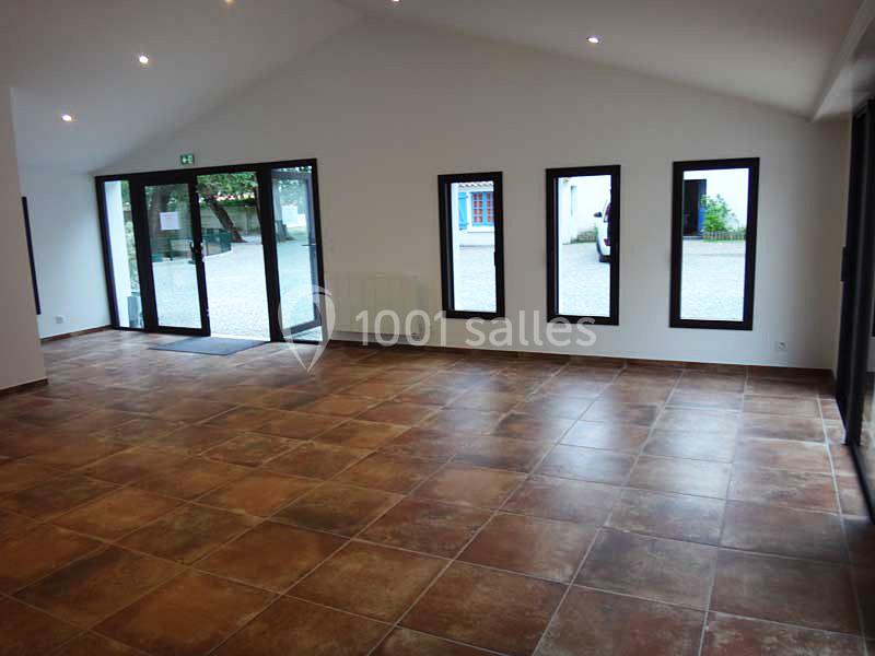 Salle vide avec carrelage marron, murs blancs, grandes fenêtres et porte vitrée donnant sur une cour extérieure.