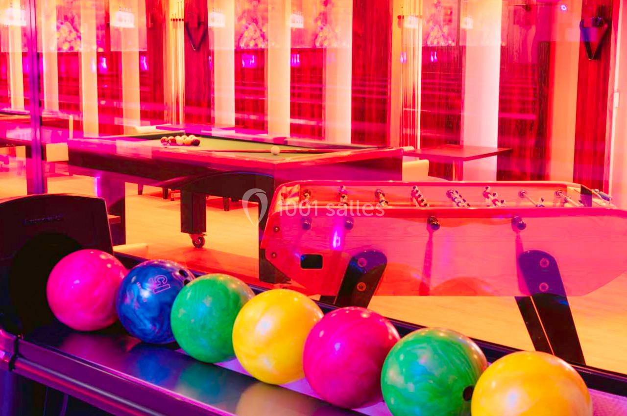 Six boules de bowling colorées alignées devant des tables de billard et un baby-foot dans une salle éclairée de lumières…