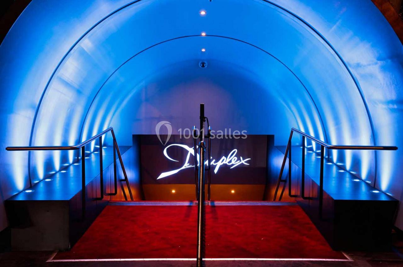 Entrée d'un espace moderne avec éclairage bleu, tapis rouge et panneau lumineux affichant un logo.