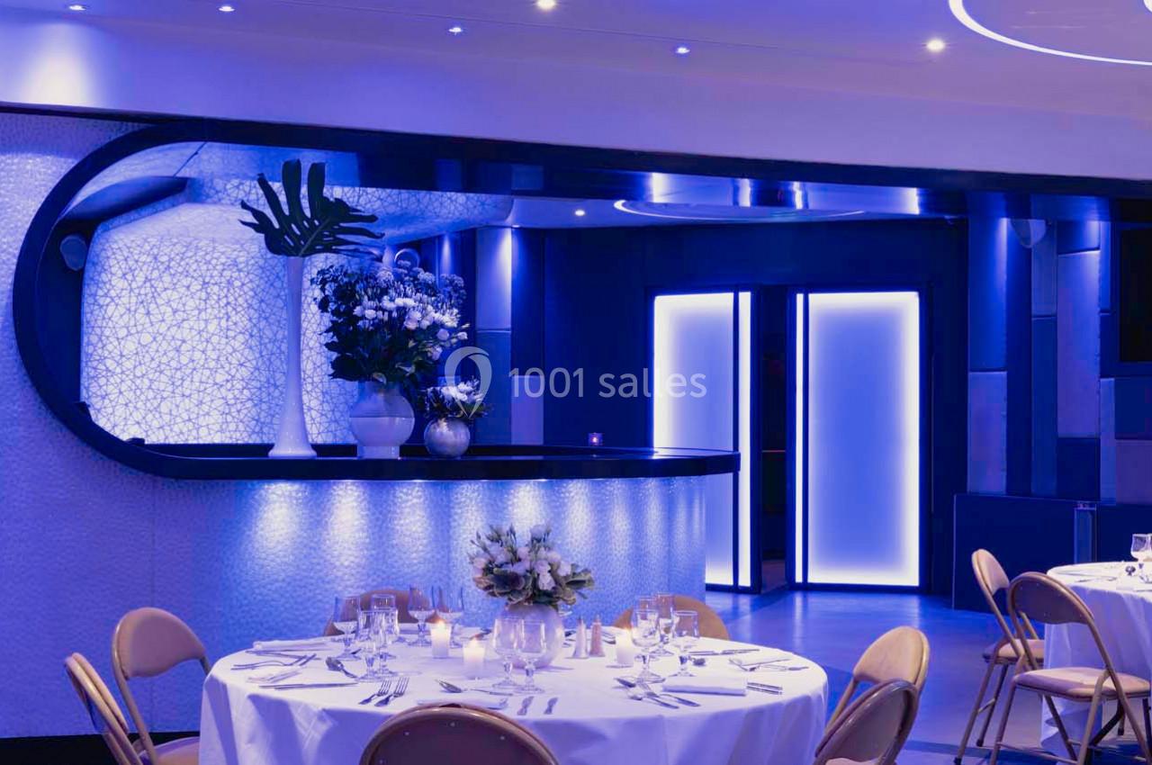 Salle de réception élégante avec tables dressées, éclairage bleu tamisé et comptoir décoré de fleurs.