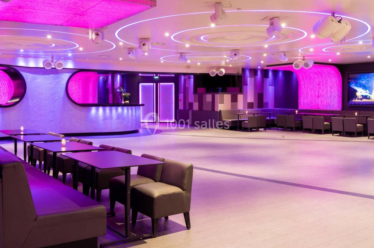 Salle moderne avec éclairage violet, tables et banquettes alignées, murs texturés et écran mural allumé.