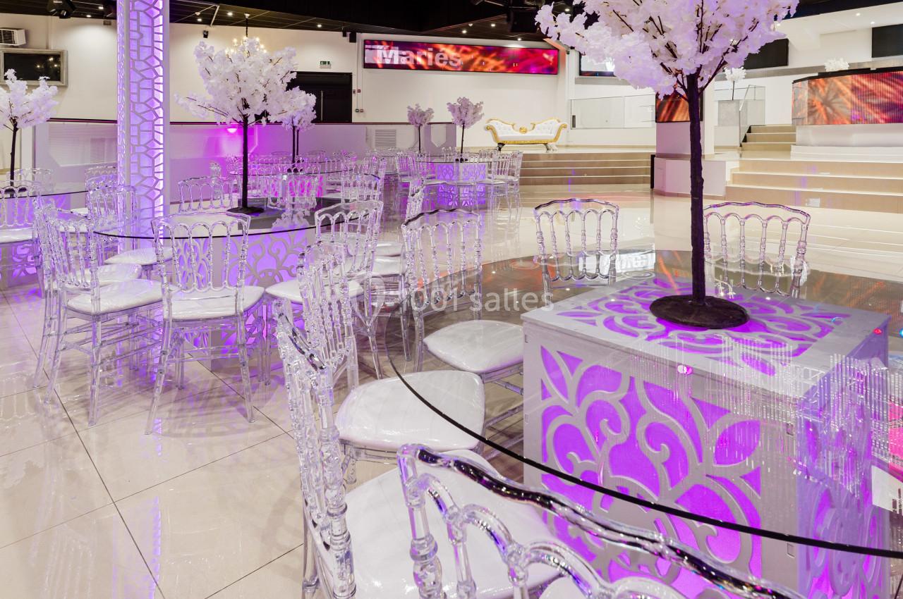 Salle de réception élégante avec tables en verre, chaises transparentes et décorations lumineuses violettes.