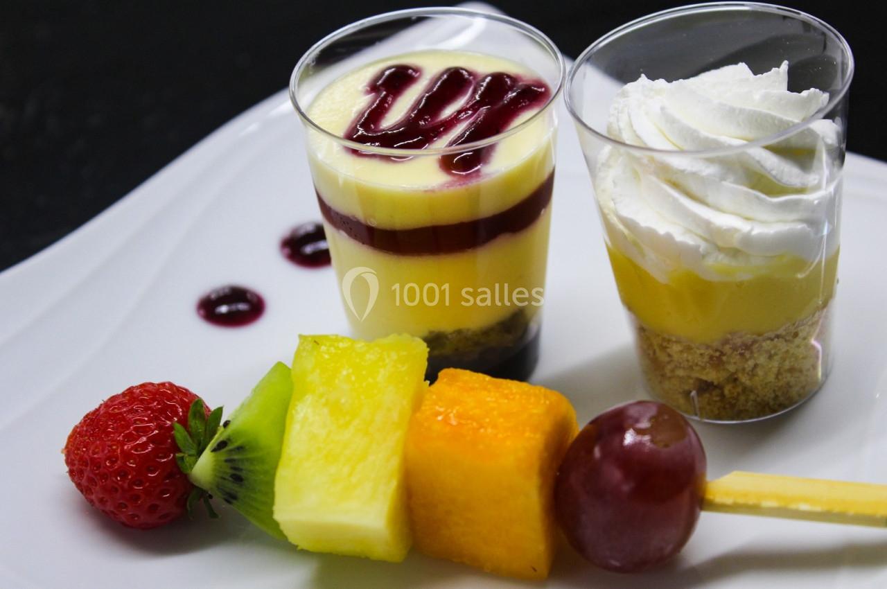 Deux verrines de dessert, l'une avec chantilly, l'autre avec coulis rouge, accompagnées d'une brochette de fruits.