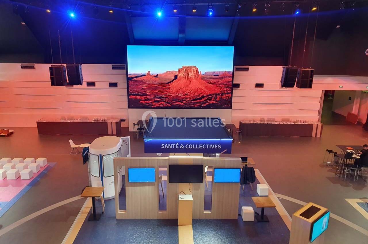 Salle d'exposition avec écran géant affichant un paysage désertique, stands en bois et espaces assis.
