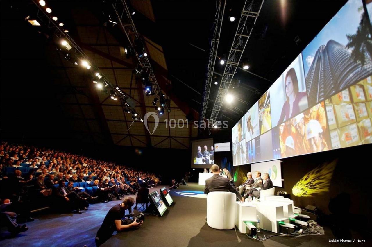 Conférence avec des intervenants sur scène, public assis dans une grande salle et écrans projetant des images variées.