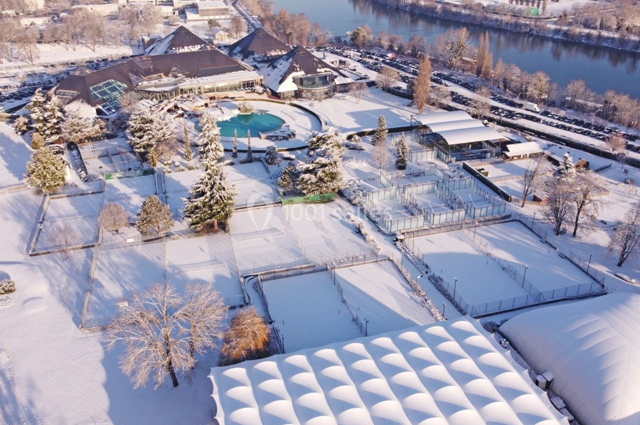 Vue aérienne d'un complexe sportif et de loisirs enneigé, avec piscine extérieure, terrains et bâtiments près d'une rivière.