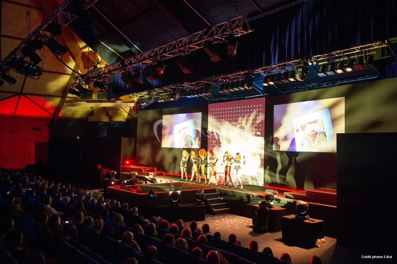 Spectacle sur scène avec des danseuses, projections visuelles et public assis dans une grande salle éclairée.