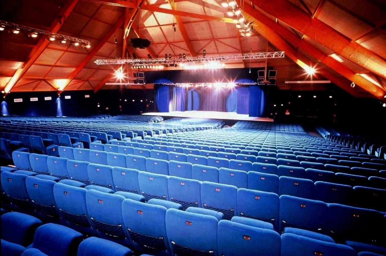 Salle de spectacle avec des rangées de sièges bleus vides, éclairée par des projecteurs sur une scène au fond.