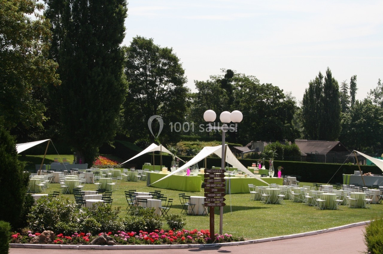 Tables et chaises disposées sur une pelouse avec des tonnelles blanches, entourées d'arbres et de parterres fleuris.