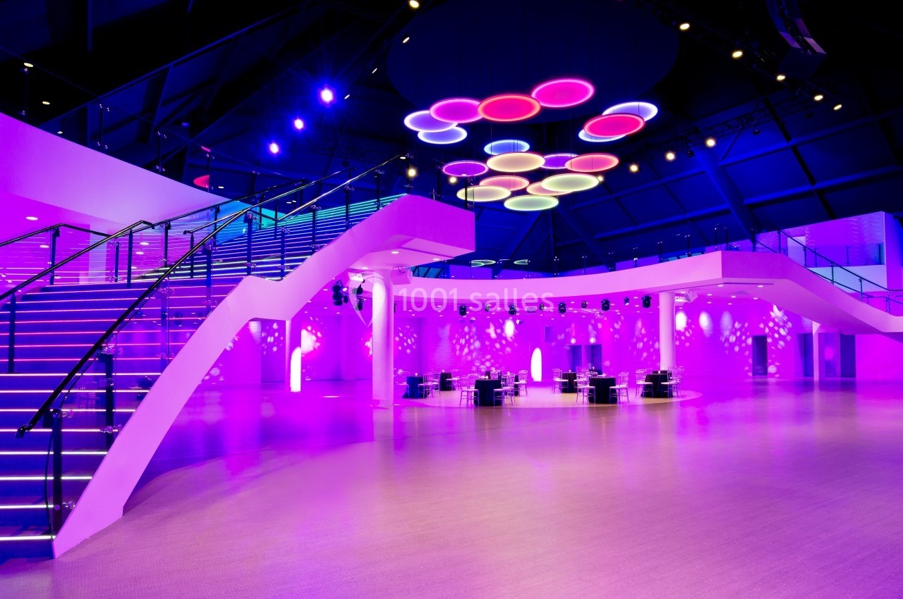 Salle moderne illuminée de lumières violettes avec escaliers, tables et plafonnier circulaire coloré.