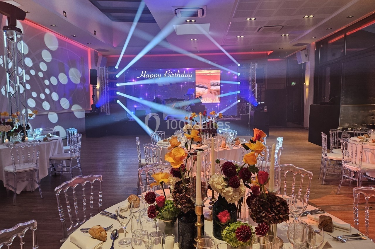 Salle de réception décorée pour un anniversaire, avec tables dressées, fleurs colorées et éclairage lumineux.