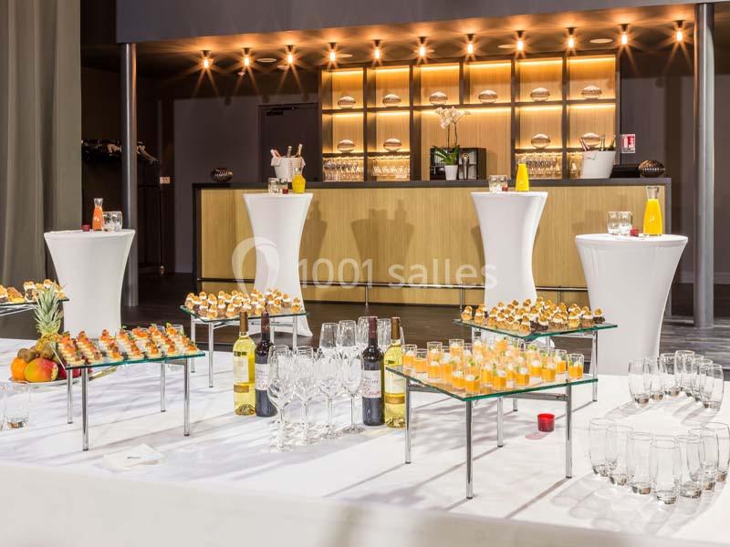 Buffet avec verrines, bouteilles de vin, jus d'orange et tables hautes dans une salle de réception moderne.