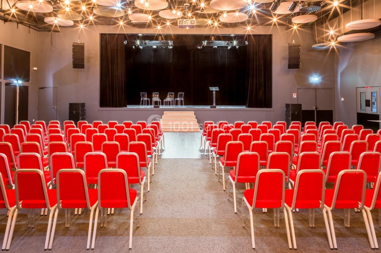 Salle de conférence avec des rangées de chaises rouges face à une scène équipée de chaises et d'un pupitre.