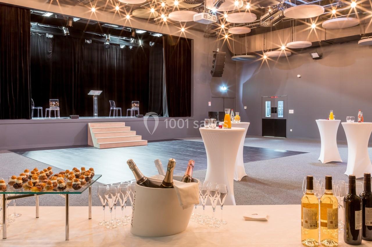 Salle de réception avec scène, tables hautes dressées et buffet comprenant boissons et amuse-bouches.