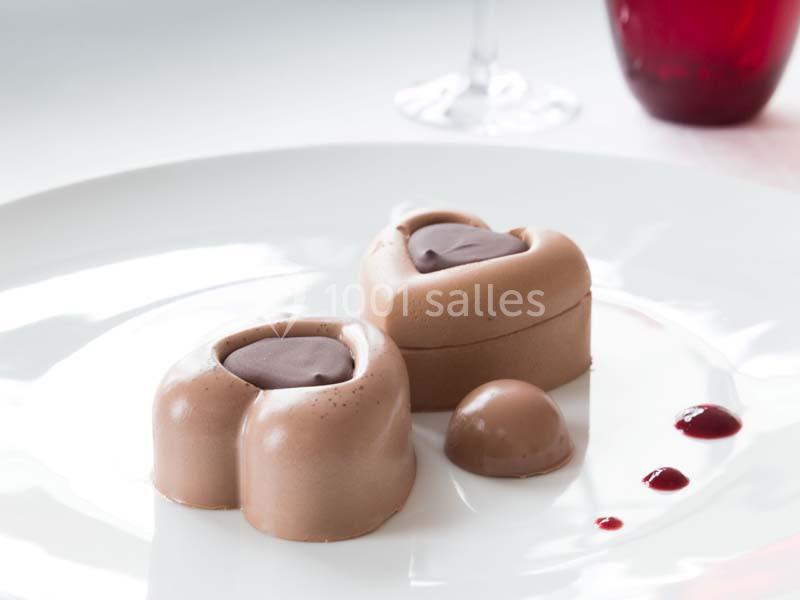 Dessert en forme de cœur au chocolat sur une assiette blanche, accompagné de coulis rouge et d'un verre en arrière-plan.