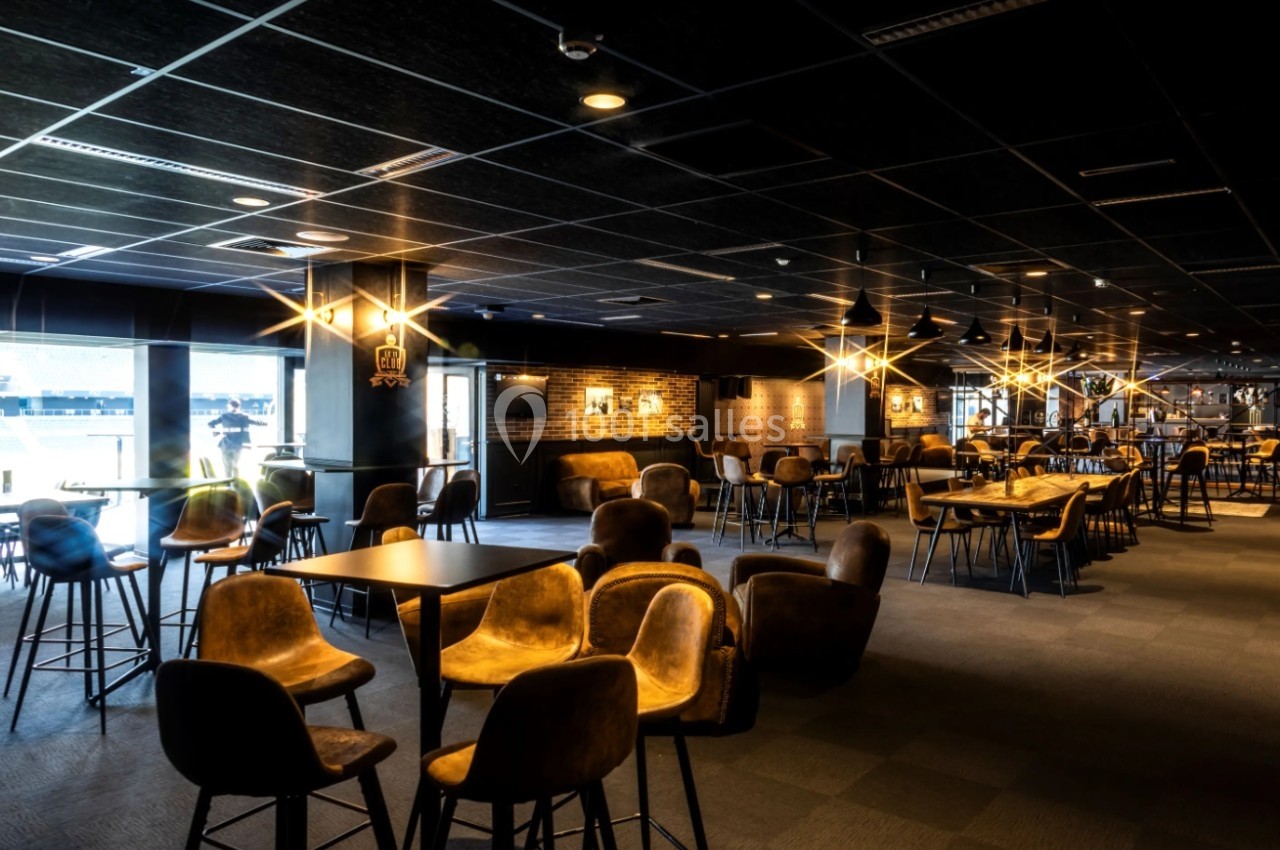 Salle moderne avec chaises en cuir, tables hautes et éclairage chaleureux dans un espace sombre et spacieux.