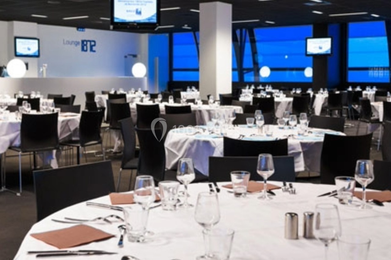 Salle de réception moderne avec tables rondes dressées, chaises noires et écrans accrochés aux murs.