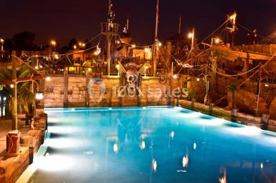 Vue nocturne d'un complexe de loisirs avec piscine illuminée, bâtiments thématiques et paysage urbain en arrière-plan.